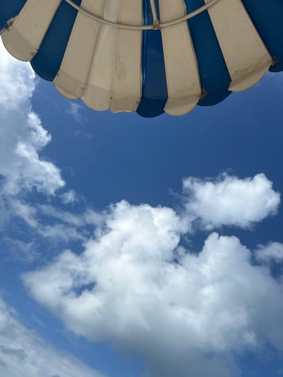 An upward-angled shot features a blue and white striped sunshade to the left, contrasted with a blue sky dotted with white cumulus clouds. The sunshade's fabric panels are visibly worn. The environment consists of fair weather conditions and a seaside location, which suggests a possible place in Le Gosier, France. There are no people visible in the image. The key objects are the sunshade and the clouds in the sky.