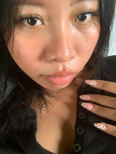 A close-up, low-angle selfie shows a person's face and hand against a black garment. The person has dark hair, dark eyes with long eyelashes, and light-pink, glossy lips. Their skin appears to have a dewy sheen. The person is wearing a gold necklace with a small, diamond-encrusted charm. Their fingernails are decorated with pink French tips and black polka dots. The person's right hand is positioned near their face, with fingers slightly curled. The background is nondescript and appears to be an indoor setting. There is no visible indication of the city of Jajag, Indonesia, or any specific environmental features that would place it there.
