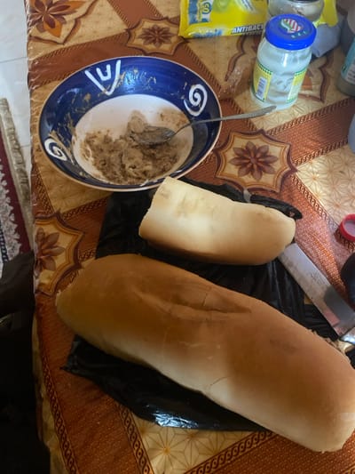 This image captures a domestic scene, likely a meal being prepared or consumed in Lakka, Sierra Leone. The primary subjects are food items and kitchenware laid out on a patterned tablecloth.

In the foreground, a large, light brown loaf of bread, resembling a baguette, rests on a crumpled black plastic bag. To its left, a smaller, cut piece of similar bread is also on the plastic bag. A metal kitchen knife with a dark handle is visible on the bag, suggesting the bread has been or will be cut.

To the left of the bread, a blue and white ceramic bowl with spiral and geometric patterns contains a brownish, pasty substance, possibly a spread or a mashed food item. A silver spoon is resting inside the bowl. Further to the left, a clear plastic jar with a blue lid and a green and yellow label is visible, containing a white substance. Next to it is a yellow plastic packet, identified by visible text as "ANTIBACTERIAL wipes" and showing the number "50". A red plastic bottle cap is also partially visible near the top right.

The items are arranged on a tablecloth featuring an ornate brown, beige, and orange pattern with star-like floral motifs and geometric borders. Part of a patterned rug or mat is visible on the floor beneath the table. The scene suggests an informal setting for a meal or food preparation. No people are visible, and the time of day is indeterminate, but the lighting suggests an indoor setting.