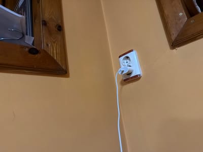 The image shows a light-orange wall with a white electrical outlet in Zlatna Panega, Bulgaria. A white electrical cord is plugged into the outlet, extending upwards. Visible on the wall are parts of two wooden frames. The scene appears to be indoors.