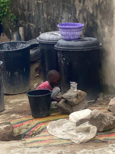 The image portrays an outdoor scene in Idon Kasa, Nigeria, against a weathered, light-colored concrete wall. A young child, possibly male, with dark skin and a shaven head, is centrally positioned, crouched low to the ground. He wears a red and white patterned shirt and light-colored trousers, looking downwards as if interacting with an object on the ground directly in front of him.
To the child's left, a black plastic bucket rests on the uneven ground. To his right, a stack of three natural rocks supports two clear plastic cups, one containing a transparent liquid. The ground beneath and around the child is a mix of dirt and concrete, partially covered by a striped mat featuring red, green, and yellow patterns. A crumpled white piece of fabric lies on this mat, near the rocks.
In the background, multiple large, black plastic storage bins with lids are visible, positioned against the concrete wall. A purple plastic basin sits atop the lid of the bin closest to the right. Faint details of vehicle tires and sparse green vegetation are discernible in the far background.