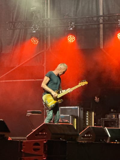 The image depicts a live musical performance on a stage at night. A male musician, appearing to be Sting, is center-frame, playing a bass guitar. He is wearing a dark t-shirt and light-colored pants. Red stage lights are prominently illuminating the scene from above and behind the performer. In the background, equipment such as speakers and lighting rigs are visible. There is a faint outline of another person in the darkness to the right of the main subject. The overall atmosphere is that of a concert, with a backdrop of dark fabric and metal trussing. The image was taken in Chiclana de la Frontera, Spain.