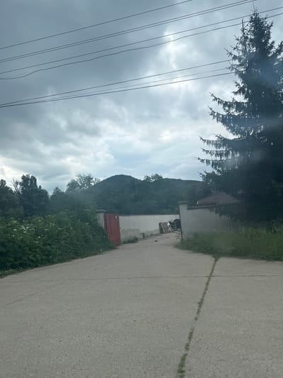 The image depicts a cloudy, overcast day in Kaleytsa, Bulgaria. A paved road leads towards a compound with a light-colored wall and a red gate. Lush green foliage and trees surround the wall, with a large coniferous tree on the right side of the frame. The sky is filled with dark, heavy clouds, suggesting an impending storm. There are no people visible in the image, and no actions or interactions are taking place. The scene appears to be a rural or semi-rural area, possibly on the outskirts of the town.