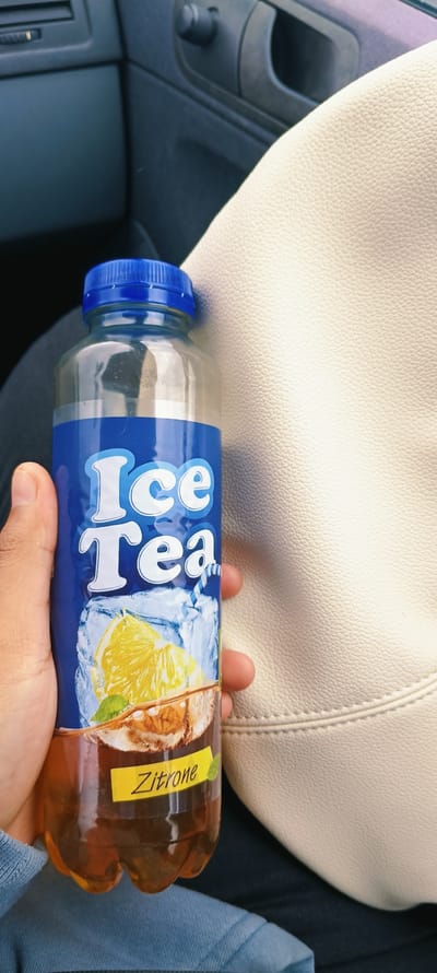 A person's hand is holding a plastic bottle of "Ice Tea" with a lemon flavor. The bottle has a blue cap and a label with white text that says "Ice Tea" over an image of ice cubes and a lemon slice. Below that, a yellow sticker reads "Zitrone," which is German for lemon. The bottle contains a brown liquid. The background shows part of a car's interior, with a dark gray dashboard and door panel, and a light-colored textured surface, possibly leather, which could be a car seat or a bag. The image appears to be taken inside a vehicle, likely in a city like Ansfelden, Austria, given the context provided in the prompt, although no explicit landmarks or street views are visible.