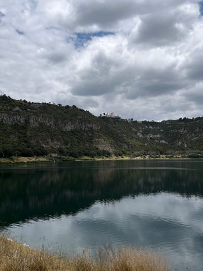 The image depicts a serene landscape featuring a calm body of water reflecting the surrounding environment. On the far bank of the water, a lush green hillside rises, covered in dense vegetation and dotted with trees. Atop the hill, a prominent building with red roofs and white walls is visible, suggesting a church or a similar structure. The sky above is overcast with a mix of dark and light gray clouds, with occasional patches of blue peeking through. The foreground shows dry, tall grass along the edge of the water. The scene appears to be the Laguna Alchichica, a crater lake located in the municipality of San Juan Atenco, Puebla, Mexico.