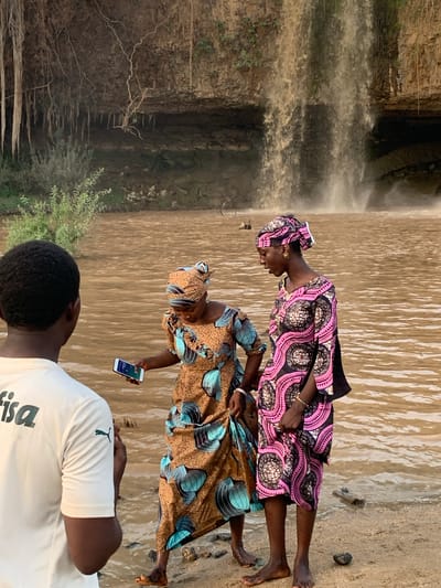 Two women and a man stand near Gurara Falls in Kukum, Nigeria. The falls are on the left. The women are wearing patterned dresses and head coverings, with the woman in the front holding a smartphone. The man is wearing a white shirt with a logo on the front. The ground appears to be sandy and wet.