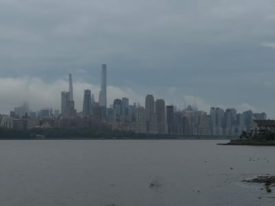This media file depicts a wide, serene view of a hazy city skyline across a body of water. The subjects are the city and the water. The city is composed of numerous skyscrapers of varying heights and architectural styles, some shrouded in mist, giving the impression of a large urban center. The water in the foreground is calm, with a few small waves rippling its surface. A few pieces of driftwood and a single bare branch are visible in the water, hinting at the natural elements interacting with the urban landscape. On the far right of the image, a house with a brown roof and white siding is situated on a grassy knoll, a sign of residential life near the city. The sky is overcast and a muted gray, suggesting a cloudy or foggy day. The overall mood is quiet and somewhat subdued, with the haze lending a dreamlike quality to the scene. The time of day appears to be daytime, but the overcast conditions prevent clear shadows or bright sunlight. The location is identified as Edgewater, USA, and the city in the distance is likely New York City, given the recognizable skyscrapers. There is no visible text within the image.