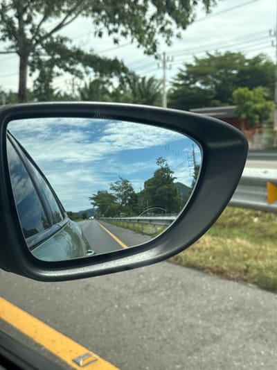 A side-view mirror of a moving vehicle reflects a road with a yellow line, green trees, and a cloudy sky. In the foreground, the asphalt road surface is visible with a yellow line marking its edge. The side-view mirror shows the reflection of the road stretching into the distance, bordered by a guardrail and lush green vegetation. The sky is a mix of blue and white clouds, suggesting a partly cloudy day. The scene appears to be set in a rural or suburban area. Based on the provided location, this scene is likely taking place in Tambon Tha Uthae, Thailand, given the tropical vegetation and the style of road infrastructure.