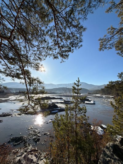 The image captures a landscape in the city of Manzherok, Russia, featuring a partially frozen river with mountain ranges in the background. The foreground includes trees with bare branches. The sky is blue, and the sun is visible, reflecting off the water. Rocks and patches of ice are present along the river's edge.