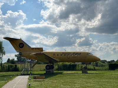 The image shows a yellow airplane on display outdoors, with the Ukrainian flag visible in the background. The plane is situated on a grassy area, with a paved walkway and steps leading up to it. The sky is filled with dramatic clouds, suggesting a partly cloudy day. There is text on the side of the airplane that reads "Yellow Plane" and "#chargenjoy". Another piece of text on a signpost reads "Вхід в літак" which translates to "Entrance to the plane." The overall scene appears to be a tourist attraction or a museum exhibit. The time of day is likely afternoon, given the lighting conditions. The image is oriented vertically, with the plane appearing upside down. The location is Zavalivka, Ukraine.