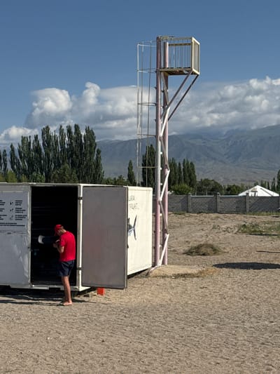 A young man wearing a red t-shirt and dark blue shorts stands with his back to the viewer, facing into an open white shipping container. He is holding a dark cylindrical object, possibly a paddle or pole, and appears to be inspecting it. The shipping container is open on one side, revealing shelves and equipment inside. The scene is outdoors on a sandy, dry ground. Behind the container is a metal tower with a caged platform at the top, painted in pink and white. In the background, there are tall green trees, a low stone wall, and mountains under a blue sky with white clouds. This appears to be a scene at a recreational area or beach, possibly near Lake Issyk-Kul in Koshkol', Kyrgyzstan, given the landscape and typical amenities for water sports.