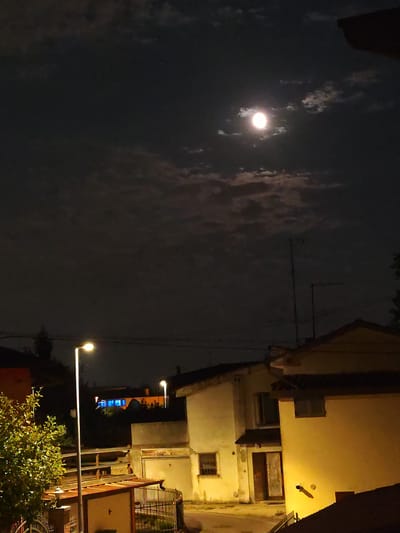 The image depicts a nighttime scene in Selvazzano Dentro, Italy. The sky is dark and partly cloudy, with a bright full moon visible in the upper center of the frame, surrounded by wispy clouds. In the foreground, there are several residential buildings. A street light illuminates a portion of the scene, casting a warm glow on a tree with green foliage in the lower left. The buildings appear to be of varying heights and colors, with some showing signs of wear. One building in the middle ground has windows with bright blue light emanating from them, suggesting occupancy. Another building, a prominent structure in the right foreground, is illuminated by artificial light, highlighting its yellow facade and roofline. The overall impression is a quiet, residential area at night, with the moon and street lights providing the primary sources of illumination. There are no people visible in the image.