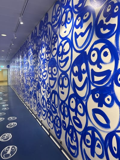 This media file depicts an interior view of a tunnel or walkway, likely in Kastrup, Denmark. The ceiling is adorned with a dense, repeating pattern of abstract blue figures painted in a bold, expressive style. These figures are circular or oval-shaped and feature stylized eyes and other facial elements, resembling a collection of cartoonish faces or characters. The painting is applied directly to a white surface, creating a striking contrast.
The walkway itself appears to be a modern structure with smooth, white walls on one side, punctuated by a series of small, recessed circular lights and a track system with adjustable spotlights. The opposite wall is a solid, deep blue, decorated with a similar repeating pattern of white abstract figures, which are smaller and more minimalist than those on the ceiling. Fluorescent lights are mounted along the top edge of the blue wall, providing illumination.
The scene is devoid of people, emphasizing the architectural and artistic elements of the space. The lighting suggests it is likely daytime or that artificial lighting is the primary source, creating a brightly lit but somewhat enclosed environment. The overall impression is one of a vibrant, art-integrated public space, possibly a transit hub or an art installation in a public area. There is no visible text in the image.
