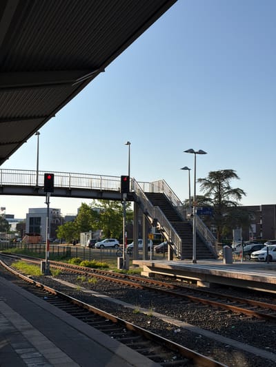 The image depicts a train station in Cleves, Germany, on a clear day with a bright blue sky. In the foreground, train tracks run parallel to a platform. To the left, a corrugated metal overhang provides shelter, casting a shadow on the scene. A pedestrian overpass, constructed with metal railings, spans the tracks. Traffic lights on the overpass are illuminated red. A metal staircase leads up to the overpass on the right side of the image. Beyond the tracks and overpass, a parking lot is visible with several cars parked. Trees and buildings are situated in the background. The overall atmosphere is one of calm and order, typical of a functional transportation hub.