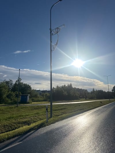 In Harkujärve, Estonia, under a bright, sunlit sky with scattered clouds, a wide, wet asphalt road reflects the brilliant sun. A tall lamppost with an ornate, globe-like fixture is positioned to the right of the road. To the left of the road, a grassy verge slopes upwards, bordered by a low curb. Further back, beyond the verge, a small, blue utility box sits beside another lamppost. Beyond that, a line of trees forms a backdrop, with several residential buildings peeking through the foliage. The overall scene is of a sun-drenched suburban or rural roadside environment.