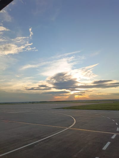 The image captures a view of an airport tarmac under a cloudy sky with the sun setting. The foreground is dominated by the asphalt surface of the runway, marked with white and yellow lines indicating pathways and boundaries. Beyond the tarmac, a strip of green grass is visible, followed by a treeline on the horizon. The sky is a mix of blue and grey clouds, with the setting sun casting a warm glow and its rays piercing through the clouds, creating dramatic lighting. The scene suggests an operational airport environment, likely at the end of the day, given the sunset and the serene atmosphere. The location is identified as Ufa Airport, Russia. There are no people or aircraft visible in the frame, focusing solely on the airport infrastructure and the sky.
