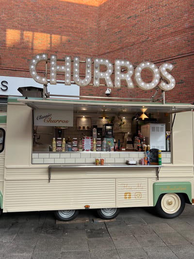A food truck specializing in churros is parked outdoors, possibly in the city of Derry/Londonderry, United Kingdom, given the architectural style of the brick building behind it and the urban ground paving. The truck is light cream and mint green, with "CHURROS" spelled out in large, illuminated marquee letters above the serving window. The serving window is open, revealing a display of various toppings, sauces, and ice cream cones. A sign inside reads "Classic Churros." On the side of the truck, "Hire Me" is written, along with the Facebook and Instagram logos, and the handle "@cullensstreetfood". The ground appears to be paved with dark, textured tiles, and the backdrop is a wall of red brick. The lighting suggests it may be late afternoon or early evening, with sunlight casting shadows on the brickwork. There are no people visible in the image.