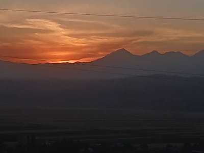 This image captures a dramatic sunset over a mountainous landscape. The sky is ablaze with vibrant orange and yellow hues, fading into a soft peach and then a dusky gray at the top. Wispy clouds are illuminated by the setting sun, creating a beautiful contrast against the darker sky.
Silhouetted against the fiery sky are the jagged peaks of mountains. They appear layered, with some ranges closer and darker than others, receding into the distance. The topmost range features sharp, prominent peaks, suggesting a rugged terrain. Below the mountains, a vast, dark expanse of land stretches out, likely a valley or plain. This area is so dim it's difficult to discern details, but hints of fields or rural structures can be vaguely made out in the lower part of the frame.
Two thin, dark horizontal lines, likely power lines, traverse the sky in the upper and middle sections of the image, providing a sense of scale and human presence within this natural setting.
The overall mood is serene and majestic, evoking the awe-inspiring beauty of nature at the close of day. The fading light suggests it is dusk. The location context provided is Taran-Bazar, Kyrgyzstan, which fits the mountainous terrain and vast open spaces depicted. There is no visible text in the image.
