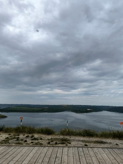 The image presents a panoramic landscape captured from an elevated position in Horaivka, Ukraine. The upper half of the frame is filled with a dense, uniform layer of gray overcast clouds, featuring a distinct dark circular spot in the upper-left quadrant. The mid-ground is occupied by a wide, calm body of dark gray water, likely a river or reservoir, which reflects the somber sky. This water feature is bordered by distant landmasses, displaying undulating terrain covered by a mix of dark green dense vegetation and lighter, possibly agricultural, fields. The immediate foreground consists of a light-colored wooden deck composed of parallel planks. Beyond the deck, a strip of sparse, dry grass and earth precedes the water's edge. On the left side of the foreground, a brown rectangular sign is partially visible, mounted on a pole with alternating green and yellow stripes. A second, identical green and yellow striped pole is present on the right foreground. No individuals are depicted in the scene.