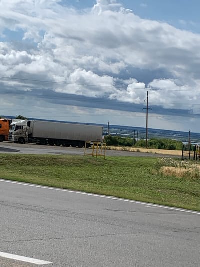 The image shows a gray asphalt road in the foreground. Beyond the road is a strip of green grass, followed by a dirt or gravel area with some yellow barriers. Further back, there are several large trucks parked side-by-side. The trucks are white and appear to be semi-trailer trucks. In the background, there is a landscape with green trees and some water, possibly a river or lake, under a cloudy sky. There are utility poles and wires visible. The scene appears to be in a rural or semi-rural area, consistent with the outskirts of Soroch'i Gory, Russia. There are no people visible in the image.