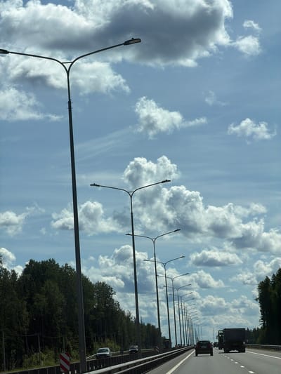 The image depicts a highway scene under a cloudy sky, likely in or near Brilliant, Russia, given the context. A series of tall lampposts line the right side of the road, extending into the distance. Several vehicles are visible on the multi-lane highway, including cars and trucks. The road is bordered by a forest on the left and right sides, with dense green trees forming the backdrop. The sky is a vibrant blue, interspersed with large, fluffy white clouds. The overall impression is that of a typical highway journey on a day with dynamic cloud formations.