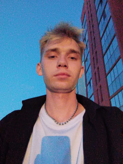The media file is a close-up, selfie-style photograph of a young man standing outdoors in front of a modern building. The man has light-colored, spiky hair and is looking directly at the camera with a neutral expression. He is wearing a white t-shirt and a dark jacket, accessorized with a silver chain necklace. The background features a tall building with numerous windows and a clear blue sky. The lighting suggests it is either early morning or late evening, possibly dusk, due to the blueish hue of the sky and the way the building's windows reflect the light. The overall setting appears to be urban. There is no visible text in the image. The location context is Zapadny, Russia.