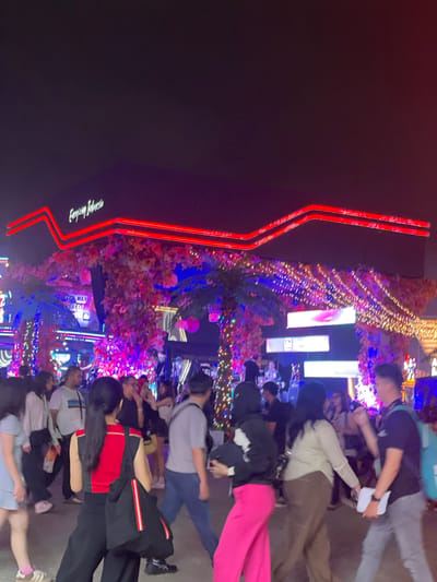 The image depicts a nighttime scene in Salembaran Jati, Indonesia, showcasing a crowded outdoor area illuminated with decorative lights. A prominent red neon sign for "Emporium Indonesia" is visible on a dark building facade to the left. The street is filled with people, some walking, others standing, suggesting a busy public space. The setting is festive, with trees and structures adorned with strings of lights in various colors, creating a vibrant atmosphere. The attire of the individuals includes casual clothing, such as t-shirts, pants, and jackets. The lighting is bright and colorful, contributing to the lively ambiance.