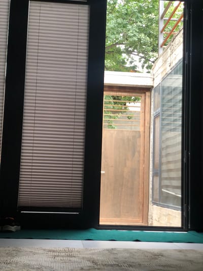 An interior view from a room with blinds looks out onto a courtyard with a brown metal door and a window covered with blinds. A tree with green leaves is visible outside. The floor inside is covered with a rug and a green mat. A pair of athletic shoes are on the mat near the blinds.