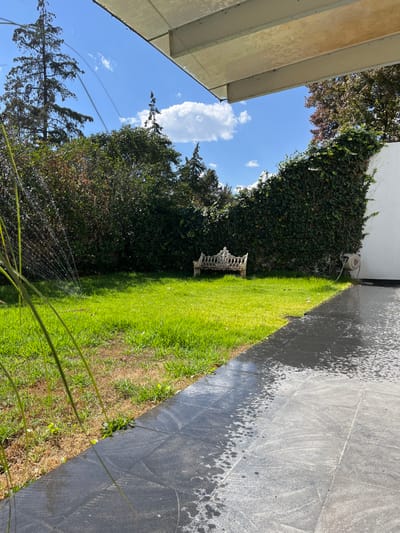 An outdoor scene features a green lawn extending from the bottom right to the upper left. A decorative white bench sits on the grass near a vine-covered wall. To the right of the lawn, a dark, tiled surface is visible. A sprinkler is in operation, with water droplets creating a hazy effect in the foreground. In the sky, there's a prominent white cloud against a blue backdrop. Trees surround the space, and a portion of a building's roof is visible in the upper left corner.