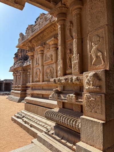 The image shows the underside of a stone structure, likely a temple, in Kamalapura, India. The structure consists of multiple layers of intricately carved stone beams and pillars. The carvings depict various figures, floral patterns, and geometric designs. The stone is a light brown color. The sky is visible in the lower left corner, indicating that the photograph was taken during the day. The overall perspective suggests the photo was taken from a low angle, looking up at the structure.