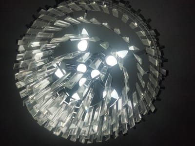 The image shows a close-up, upward-facing view of a modern chandelier. The chandelier is circular and features multiple tiers of clear, faceted crystal prisms. These prisms are attached to a metallic frame, creating a tiered, cascading effect. Within the structure of the chandelier, several bright, round light bulbs are visible, illuminating the crystals and casting reflections. The background is a dark, solid color, likely the ceiling, which makes the illuminated chandelier stand out prominently. The lighting suggests the image was taken indoors, possibly at night or in a dimly lit room, where the chandelier is the primary light source. The detailed focus on the crystals highlights their prismatic qualities, with hints of rainbow refractions visible in some areas. There is no text present in the image.