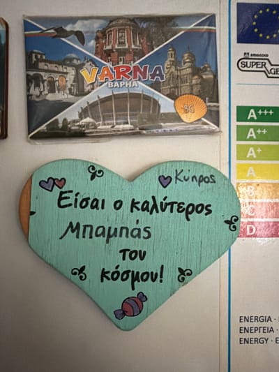 The image shows a close-up view of a refrigerator door with several items attached. On the top left, there is a postcard depicting scenes of Varna, Bulgaria, including buildings and a stadium. Below the postcard, a light blue wooden heart-shaped ornament is prominently displayed. The heart is adorned with black lettering in Greek, which appears to say "Είσαι ο καλύτερος Μπαμπάς του κόσμου!" (You are the best Dad in the world!). There are also small drawings on the heart, including two purple and pink hearts, a scroll-like design, a purple heart with "Κύπρος" (Cyprus) written next to it, and a drawing of a wrapped candy. To the right of the heart ornament, a vertical energy efficiency label is partially visible, showing color-coded categories like A+++, A++, A+, A, B, C, and D, followed by text that includes "ENERGIA" and "ENERGY". The overall scene suggests a domestic setting, likely a kitchen, where personal mementos and informational labels are displayed on a refrigerator. The presence of the word "Cyprus" on the heart ornament suggests a connection to Dromolaxia - Meneou Municipality, Cyprus, as a place of origin or significance.