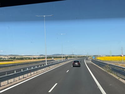 The image depicts a view from a vehicle traveling on a highway near Prolesha, Bulgaria. The highway is asphalt with white lane markings. A dark-colored SUV is in the right lane, ahead of the viewer's vehicle. On the left side of the highway, there is a metal guardrail. Beyond the guardrail, there is a field of yellow sunflowers. In the background, there are rolling hills and some scattered buildings. The sky is clear and blue. Several tall lampposts line the highway. Traffic signs are visible, including a blue circular sign with a white arrow pointing straight ahead, indicating a one-way street, and a triangular yield sign. Power lines and towers are also visible in the distance. The scene appears to be during daylight, likely late morning or early afternoon, given the brightness and the long shadows.