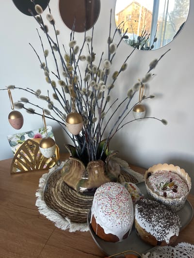 The image depicts an Easter still life scene. The setting appears to be indoors, likely on a wooden table, based on the wood grain visible. The composition features a bouquet of pussy willow branches with decorative gold-painted eggs hanging from them. There are three Easter cakes, decorated with white icing and colorful sprinkles, arranged on a dark gray plate placed on a woven placemat. A round mirror is visible in the upper left corner, reflecting the light. A decorative gold fan-shaped object and a floral-patterned item are positioned at the bottom of the image. The overall scene conveys a festive, Easter-themed atmosphere. No specific location can be pinpointed; the presence of Mežezeri, Latvia, can not be verified.