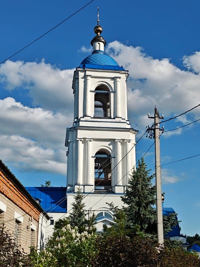 A white, multi-tiered bell tower with a blue dome and gold spire stands tall against a bright blue sky with puffy white clouds. The structure is partially obscured by trees and bushes in the foreground. An electrical pole with wires runs through the right side of the image, interrupting the view of the tower. The corner of a brick building is visible to the left. The location is Мамонтово, Russia.
