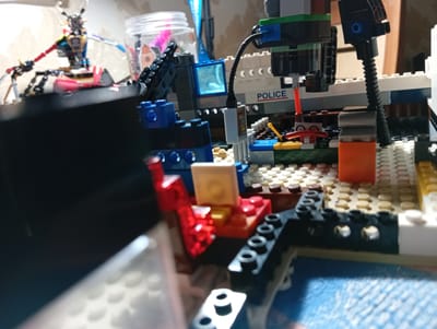 The image shows a close-up view of a Lego construction, possibly a police station, with various bricks and figures assembled. In the background, a blurred figure of a robot-like character with red and black limbs is visible, along with a transparent jar containing pink and colorful items. The Lego structure features a white and gray building with the word "POLICE" printed in red and blue. There are also blue and black Lego bricks, a red and yellow lever, and an orange Lego brick. The scene is set on a textured surface, possibly a table or floor, with a blue patterned cloth in the foreground. The lighting suggests an indoor setting, and the overall impression is of a creative Lego build. There is no identifiable information suggesting the location of this scene in Қарғалы, Kazakhstan, or any specific context beyond the Lego construction itself.