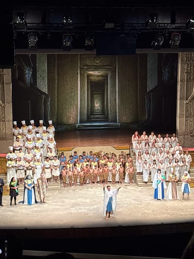 A performance of the opera Aida is taking place in the Gran Teatro Nacional in San Borja, Peru. A large cast of people, mostly dressed in ancient Egyptian-style garb, are standing in rows on the stage. The backdrop is a projection of hieroglyphic-covered walls and a deep corridor. Stage lighting equipment is visible on the left side of the frame. Audience members are also visible in the bottom right corner.