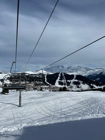 This image captures a winter alpine scene within a ski resort near Villard-sur-Doron, France, viewed from an elevated perspective. In the immediate foreground, a snow-covered slope is extensively marked with numerous ski tracks. A grey metal ski lift pylon with an attached arm is visible on the left, supporting multiple overhead cables that extend diagonally across the frame. Several empty, metallic grey ski lift chairs are suspended from these cables. The midground features a village composed of clustered buildings with various colored roofs, situated at the base of a snow-covered hill partially covered by dark green pine trees. Beyond the village, dense pine forests cover rolling terrain, intersected by visible groomed ski slopes. The distant background reveals a rugged range of snow-capped mountains under a sky displaying patches of blue amidst grey-white clouds. No individuals are present in the scene. The lighting is natural, indicative of an overcast day with some filtered sunlight.