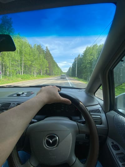 The image captures the interior of a car from the driver's perspective, with a hand on the steering wheel. The car is driving on a two-lane asphalt road through a forest. The road is lined with trees, primarily birch and pine, and there are power lines visible along the side of the road. The sky is blue with some scattered clouds. In the distance, another car can be seen driving in the same direction. The dashboard of the car is visible, including a Mazda logo on the steering wheel and a GPS device on the dashboard. The scene is consistent with a drive on a rural road in Burdakovka, Russia, during daylight hours.