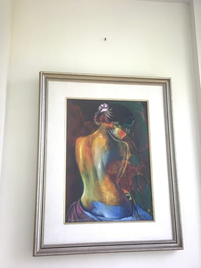 A framed painting hangs on a light-colored wall in a building located in Kota Batu, Indonesia. The painting, oriented vertically, depicts a female figure from the back, with her head and upper torso turned slightly to the viewer's left. Her back and arms are rendered with vibrant, non-naturalistic brushstrokes in yellow, orange, green, and blue hues. Her dark hair is styled in an updo, adorned with a light purple flower. A blue and purple fabric is draped around her lower back and hips. The background of the painting is dark, featuring abstract splashes of red, green, and black. A signature reading "ADENIA BALI 200" followed by an indistinct digit (possibly '3' or '9') is visible in the lower left corner of the artwork. The painting is enclosed within a silver-colored frame with a textured, beaded design on its inner and outer edges, and a wide, plain white mat board. Above the painting, on the bare wall, a single dark screw or nail head is visible, positioned slightly left of the image's vertical center. A smaller, faint dark mark is present to the immediate left of the screw/nail. The wall itself is a plain, off-white or cream color. The scene is evenly lit.