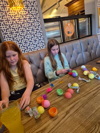 Here is a detailed description of the image:
**Scene:** An indoor setting, likely a restaurant or pub, with two young girls at a wooden table.
**People:**
* Two girls: One has light brown hair and is wearing a light blue sweater. The other has red hair and is wearing a light yellow shirt. They appear to be related, possibly sisters.
**Objects:**
* Plastic Easter eggs of various colors (pink, yellow, orange, green, purple, white).
* Small toys or candies, some still wrapped.
* A large pitcher of a yellow beverage, possibly lemonade or juice.
* A brown leather upholstered banquette or booth seating.
* Decorative wall with a repeating geometric pattern in blue and white.
* A glass window panel with frosted glass.
* A sign with orange background is attached to the glass window panel saying "DOWNLOAD THE GREENE KING APP TO ORDER & PAY".
**Actions:**
* The girls are playing with the plastic Easter eggs and their contents, possibly sorting or organizing them.
* The girl with the red hair is reaching towards or pouring from the pitcher of yellow beverage.
**Interactions:**
* The girls appear engaged in a shared activity, possibly involving Easter-themed toys and treats.
**Environment:**
* The background shows elements of a commercial establishment, such as the patterned wall, the window, and the sign.
**Location Context:** The sign mentioning "GREENE KING APP" suggests this is a Greene King pub or restaurant, which is a popular chain in the United Kingdom. This image was taken in Breckland District, United Kingdom.