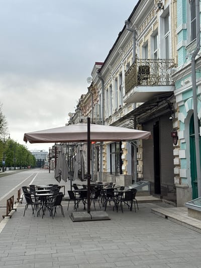 The image depicts a street scene in Akhieli, Georgia, featuring a row of closed or unoccupied outdoor cafe seating areas along a paved walkway. The building façades on the left exhibit ornate architectural details with decorative balconies and cornices. Parasols are folded and secured, and chairs are stacked. No people are visible in the immediate foreground. A street with trees lines the walkway further down the image. Weather conditions appear overcast with a gray sky.
