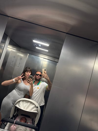 A man, a woman, and a baby are in an elevator, possibly in Pomezia, Italy. The woman holds up two fingers in a peace sign, while the man holds a phone taking a picture of their reflection in the elevator's metal walls. The baby is in a stroller. Both the man and the woman are using cell phones, and the woman is wearing a watch.