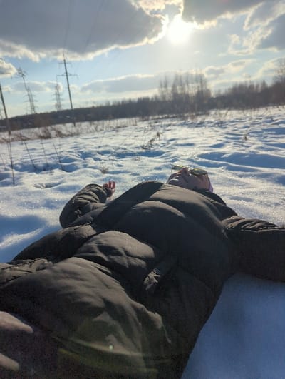 The scene depicts an individual lying supine in a snow-covered field in Podolino, Russia. The person is wearing a dark, possibly black, puffy winter jacket and has sunglasses resting on their forehead. Their arms are extended slightly to their sides. The ground is covered in textured white snow, from which sparse, dry vegetation stalks protrude in the mid-ground. In the background, multiple power line towers with overhead lines are visible, leading towards a distant treeline. The sky is blue with scattered white clouds. The sun is prominent in the upper right quadrant, emitting a bright glare and lens flare, indicating a clear, sunny winter day.