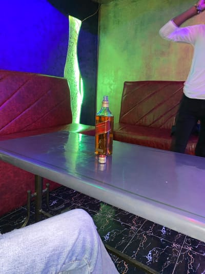 A bottle of Johnnie Walker Red Label lies horizontally on a bar top. One person sits at the bar, wearing blue jeans, while another stands nearby. The bar area features red patterned seating, a blue-lit section, and a wall decorated with black and white marble-like tiles. Green lighting casts an ambient glow.