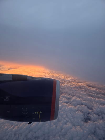 The image is an aerial view taken from an airplane window, showcasing a dramatic sunset above a sea of clouds. The foreground is dominated by the blue and gray casing of an airplane engine, with a red and white stripe on its edge. Numerous labels and warnings are visible on the engine casing. The horizon is ablaze with a vibrant orange and yellow sunset, casting a warm glow on the fluffy, white clouds that stretch out below. The sky above the clouds transitions into a soft gray, with hints of purple and blue. The scene is serene and majestic, highlighting the beauty of nature from a high altitude. The location is not specified in the image.
