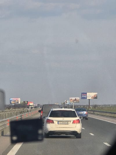 The scene depicts a multi-lane asphalt highway in Haskovo Province, Bulgaria, likely near Opalchenets, under an overcast or hazy sky. The perspective is from within a vehicle traveling on the road. In the immediate foreground, a dark, out-of-focus object, possibly part of the dashboard or an electronic device, obscures the lower left portion of the image.
The central subject is a white Mercedes-Benz station wagon, license plate "X 2434 KM", positioned in the rightmost lane of traffic, facing away from the viewer. Directly in front of the Mercedes is a dark grey or black van or SUV. Further ahead in the adjacent right lane, a blue sedan is visible. Multiple other vehicles, including a white truck, are present in the distance across various lanes, indicating active traffic.
The highway is bordered on the left by a metal crash barrier and on the right by a similar barrier separating the road from an open, flat landscape that extends to the horizon. Numerous advertising billboards are erected along both sides of the highway in the mid-distance. No individuals are visibly present outside of vehicles. The sky is a uniform light grey, devoid of distinct cloud formations.