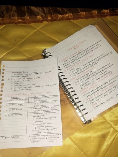 The image shows two notebooks on a yellow, quilted fabric surface. The notebook on the left is a loose-leaf paper with handwritten notes in Indonesian. The notebook on the right is a spiral-bound notebook with more handwritten notes. The notes appear to be related to legal matters, specifically the structure and development of religious courts in Indonesia.

The left notebook contains a table with "Peraturan" (Regulations) and "Kewenangan" (Authority) as columns. It lists various laws and regulations, such as UU No. 7/1989 and UU No. 50/2009, along with descriptions of their authority.

The right notebook has headings such as "Susunan Badan Peradilan Agama" (Structure of the Religious Court Body), "Pengertian Peradilan Agama" (Definition of Religious Court), and "Perkembangan Lingkungan Peradilan Agama" (Development of the Religious Court Environment). It also mentions "Kedudukan di Indonesia" (Position in Indonesia) and "Susunan Badan Peradilan Agama" again, followed by points about their functions.

The background fabric suggests an indoor setting, possibly a home or a study area. There are no people visible in the image. The lighting is dim, and there is a slight shadow cast on the notebooks. The location of Labissa, Indonesia, is not explicitly indicated in the image itself, and no elements in the scene provide clues to that specific geographical context. The image is a still shot, showing the static arrangement of the notebooks and the fabric.