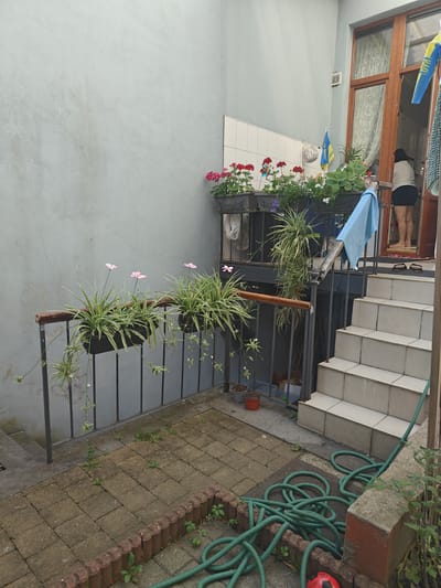 The image depicts a narrow outdoor space with a light blue-grey textured wall on the left. In the foreground, a paved area with interlocking square bricks shows some weeds growing in the gaps. A decorative brick border lines the edge of the paved area. A coiled green garden hose lies on the paving stones. To the right, a set of light-colored tiled stairs leads upwards towards an open doorway.
On a metal railing running along the paved area, several black planter boxes contain green, leafy plants, some with delicate pink flowers. Above this railing, another set of planter boxes is visible, filled with a variety of flowering plants in shades of red and pink. Behind these, a section of white tiled wall is present.
The doorway at the top of the stairs reveals an interior with kitchen cabinetry. A woman wearing a light-colored top and dark shorts is standing in the doorway, facing away from the camera. A blue towel is draped over the railing near the top of the stairs. Two Ukrainian flags are visible, one larger and blue and yellow, attached to the railing near the doorway, and a smaller one on a pole behind the upper planter boxes.
The scene appears to be a small courtyard or patio area adjacent to a residential building. The overall impression is one of a lived-in, somewhat cluttered outdoor space. The provided location information suggests this is in Saint-Josse-ten-Noode - Sint-Joost-ten-Node, Belgium.