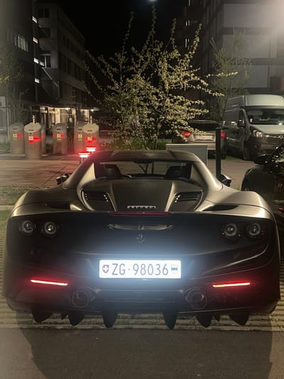 The image captures a dark-colored Ferrari sports car, likely matte grey or black, viewed from its rear in an outdoor setting at night. The car is the primary subject, displaying distinctive round taillights, a long red light strip above the license plate, and quad exhaust pipes integrated into a prominent rear diffuser. A "Ferrari" badge is visible on the trunk lid. The Swiss license plate "ZG-98036" is brightly illuminated, indicating the canton of Zug.
In the background to the left, a vibrant tree or large bush with light-colored, possibly flowering or new spring leaves, stands out against the dark sky, illuminated by ambient light. Further back, multi-story buildings with some illuminated windows suggest an urban or suburban environment.
Other vehicles are partially visible in the background; to the upper right, a white van is parked, and below it, another car displays a Swiss license plate "LU-5675704" (Luzern). Along the left side of the frame, several light-colored, cylindrical structures resembling bollards or charging stations are visible, each emitting a red glow.
The scene depicts a static moment with no overt activity, suggesting the vehicles are parked. The artificial lighting from the car's lights, the bollards, and building windows highlights the nighttime setting. The ground appears to be asphalt with some grassy pavers.